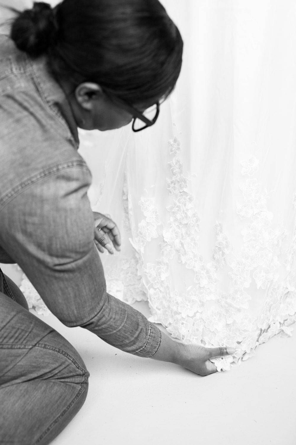 A black and white photo of the designer Kate Edmondson adjusting Lace at the hem of wedding gown,