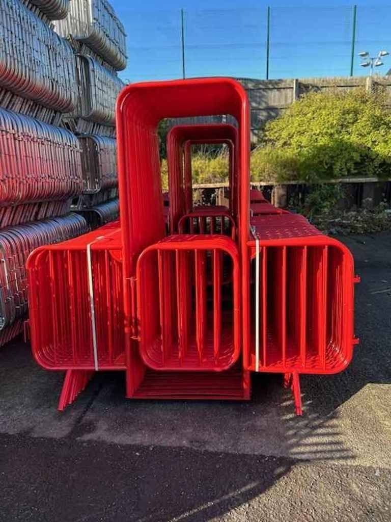 Red powder-coated crowd control barriers stored in yard at Tempfence UK with galvanised Heras fencing panels in background