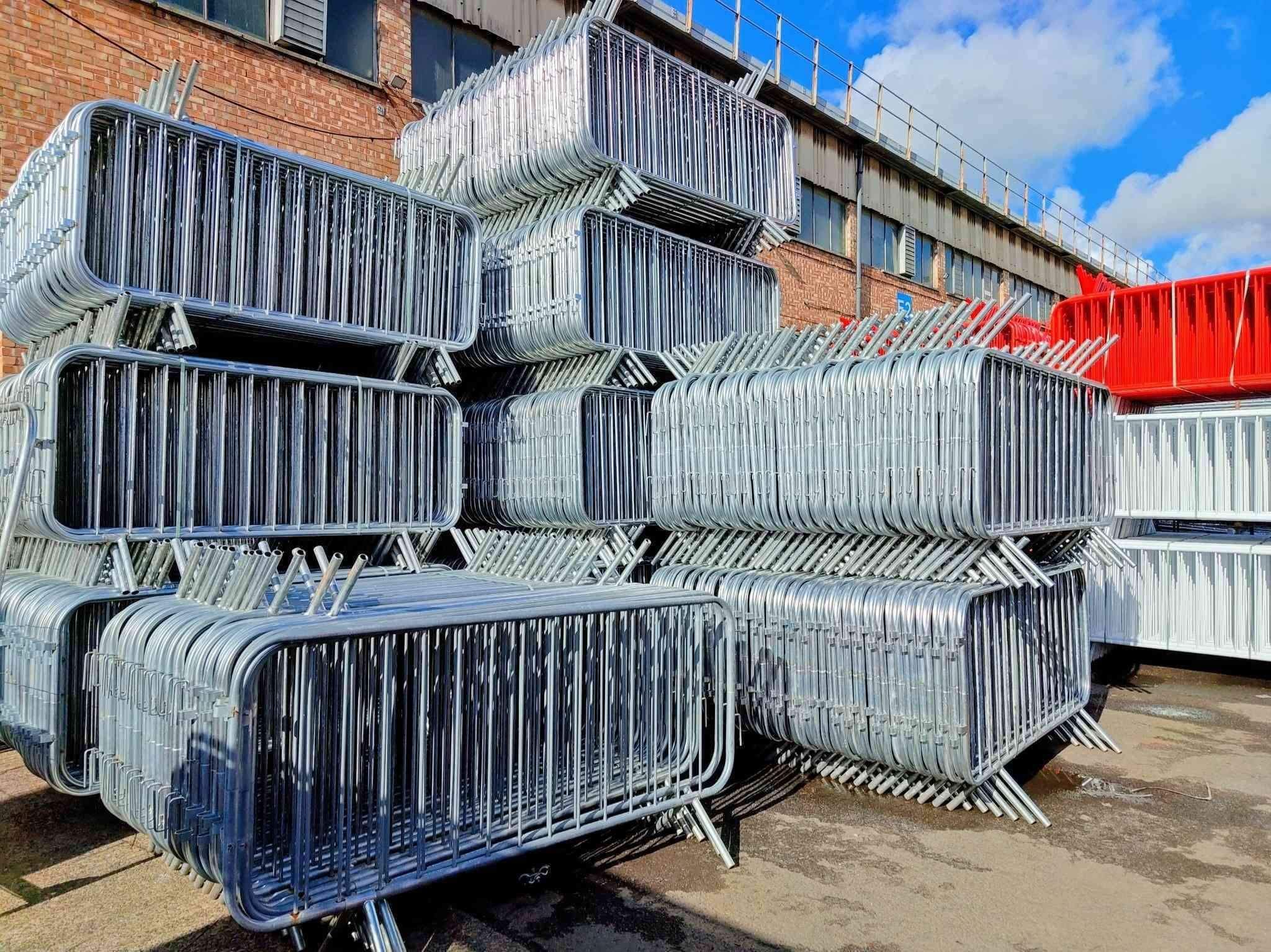 Large stock of galvanised and red crowd control barriers stacked at Tempfence UK yard, heavy-duty barriers ready for fast nationwide delivery.